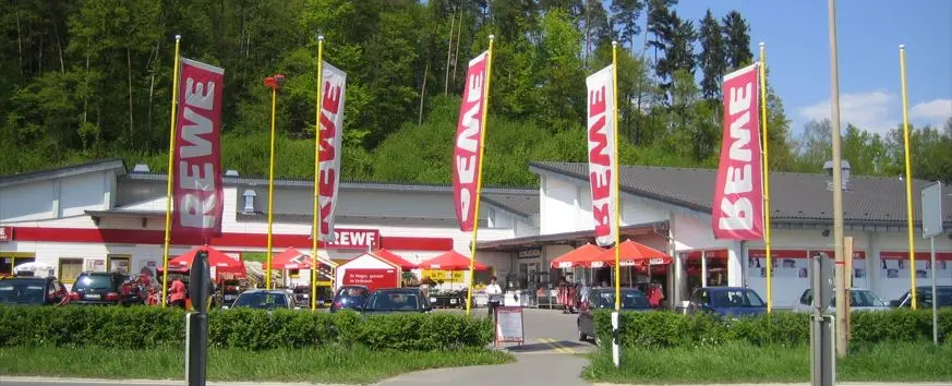 REWE supermarket in Altenkunstadt with red flags, parked cars and a green hillside behind the store.