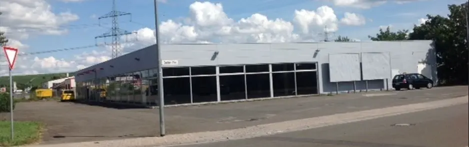 Ein langes graues Gewerbegebäude am Straßenrand in Bad Kreuznach mit großen Fensterfronten, geparktem Auto, Verkehrsschild und sonnigem Himmel.