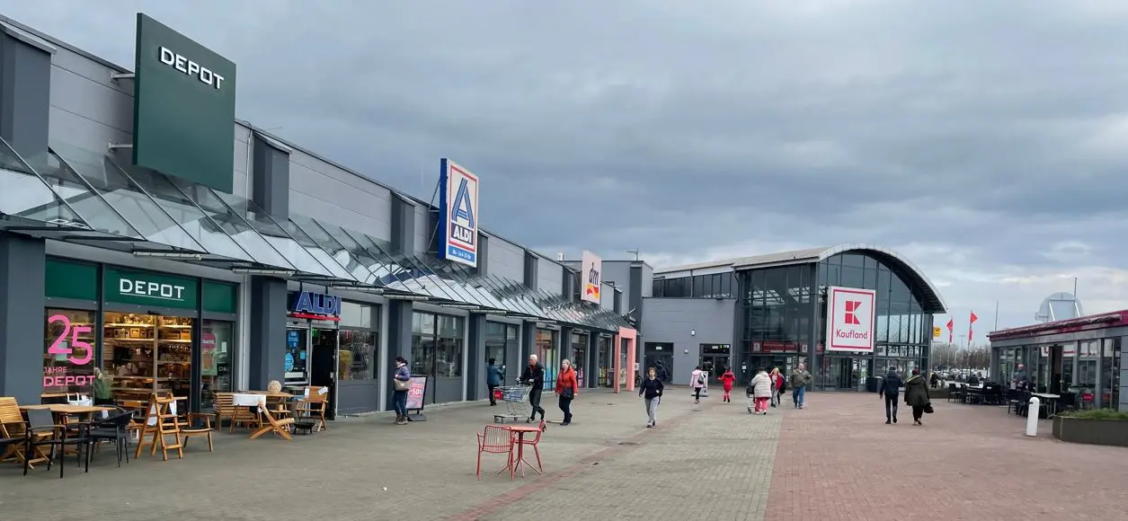 Vor einem Gewerbezentrum in Bentwisch stehen die Ladenfronten von Depot, Aldi und Kaufland unter einem grauen Himmel, während Menschen über den Platz gehen.