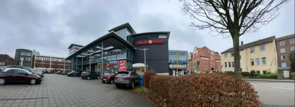 Vor dem Mediamarkt in Nienburg-Weser steht ein modernes Einkaufsgebäude mit Glasfassade, geparkten Autos und umliegenden Wohnhäusern.