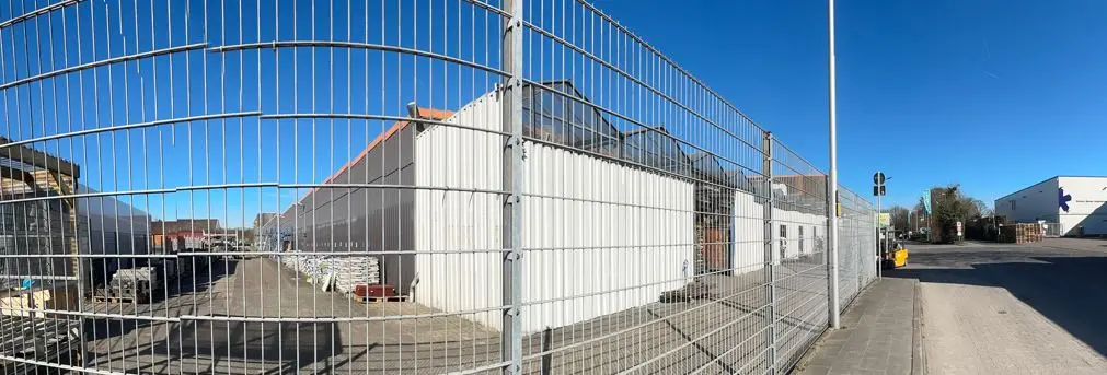 Ein großflächiges Gewerbegebäude hinter einem hohen Metallzaun an einer Straße in Straelen, mit gepflastertem Zugang und blauem Himmel.