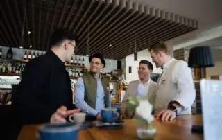 Vier Männer in einem modernen Café, die an einem Tisch stehen und miteinander sprechen, während Getränke auf dem Tisch stehen.