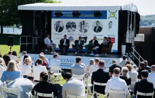 Paneldiskussion mit mehreren Sprechern auf einer Bühne vor Publikum während einer Veranstaltung im Freien.