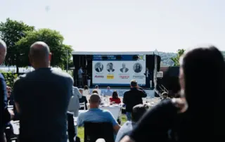 Menschen bei einer Veranstaltung im Freien, mit einer Bühne im Hintergrund, auf der Präsentationen stattfinden.