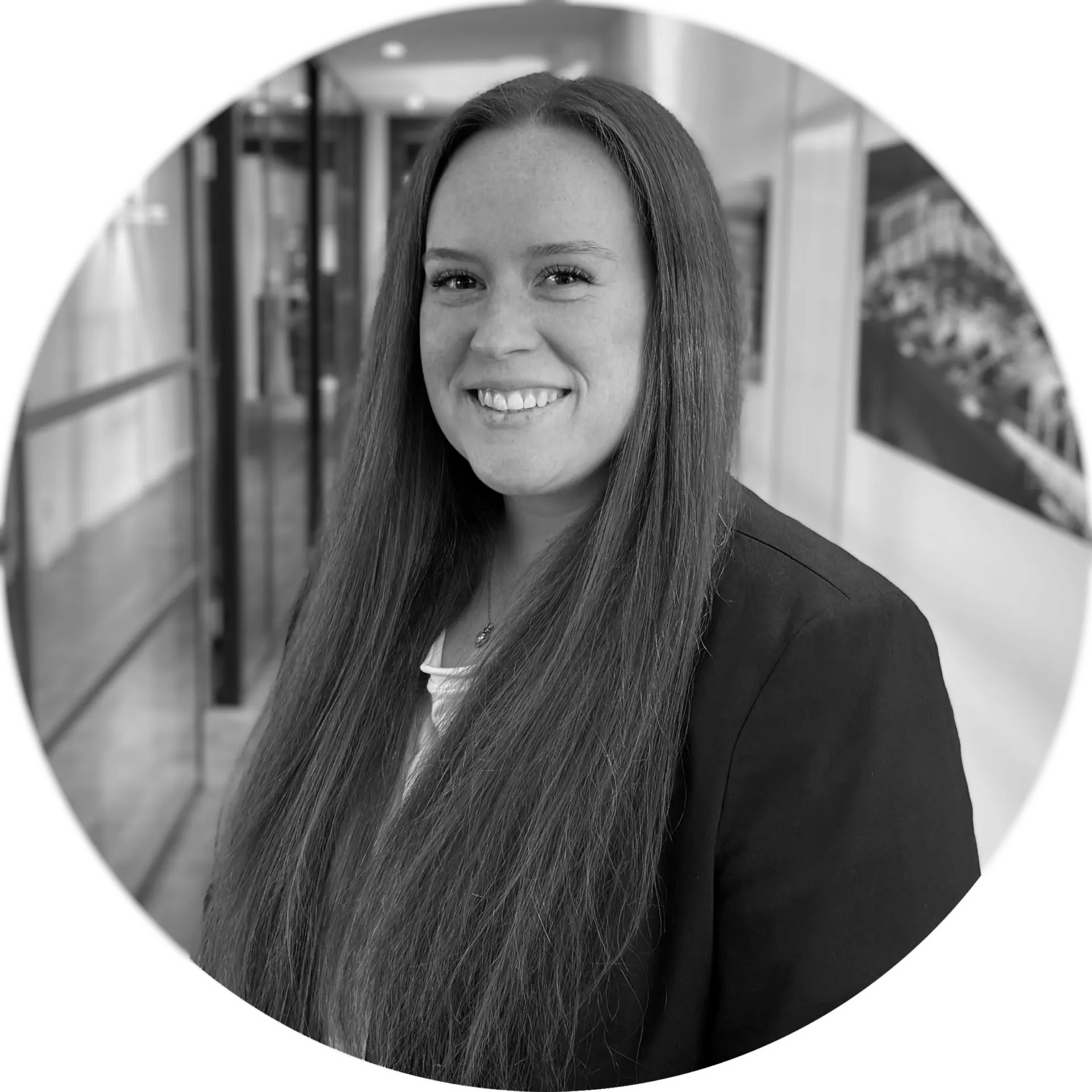 Porträt einer Frau mit langen Haaren, die in einem professionellen Outfit lächelt, vor einem modernen Hintergrund.