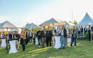 Beim Retailer Meeting am Möhnesee stehen Gäste auf der Wiese, tauschen sich unter Sonnenschirmen aus und halten Getränke in der Hand.