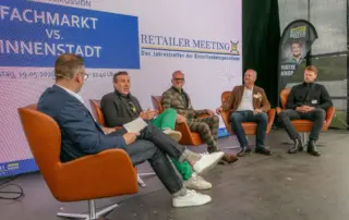 Auf der Bühne diskutieren fünf Teilnehmer beim Retailer Meeting über Fachmarkt vs. Innenstadt vor großem Eventbanner und Matze-Knop-Fahne.