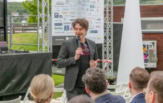 Beim Retailer Meeting am Möhnesee spricht ein Mann mit Mikrofon vor sitzenden Gästen auf einer Outdoor-Bühne mit Sponsorentafel und Blick ins Grüne.