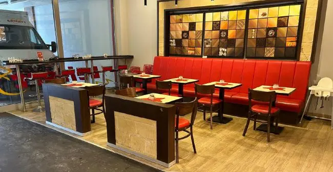 In einem Gastronomieraum in Wolfsburg stehen rote Polsterbänke, kleine Tische und Holzstühle vor einer bunten Fliesenwand und Glasfront.